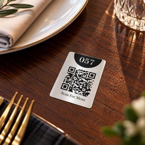 Könnte beinhalten: Ein rechteckiges Schild aus gebürstetem silberfarbenem Metall mit einem schwarzen Halbkreis oben, der die weiße Zahl 057 zeigt. Unter der Zahl befindet sich ein schwarzer QR-Code und der Text Scan For Menu in schwarzer Schrift auf der Oberfläche.