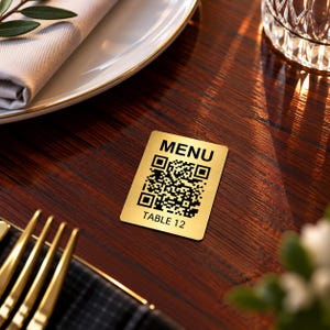 May include: A rectangular gold-colored metal sign with rounded corners featuring the word MENU in black text at the top, a central black QR code, and the text TABLE 12 in black at the bottom, designed for restaurant table identification.