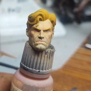 May include: A miniature sculpted head with wavy yellow hair and a stern facial expression, featuring a small red mark on the cheek. The head is mounted on top of a cylindrical paint bottle cap labeled 140 US DESERT SAND, showing detailed paintwork.