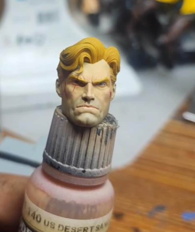 May include: A miniature sculpted head with wavy yellow hair and a stern facial expression, featuring a small red mark on the cheek. The head is mounted on top of a cylindrical paint bottle cap labeled 140 US DESERT SAND, showing detailed paintwork.