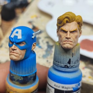 May include: Two miniature painted heads mounted on paint bottles. One head features a blue mask with a white letter A and small wings, while the other head shows a person with blonde hair and a serious expression. Both have detailed facial features.