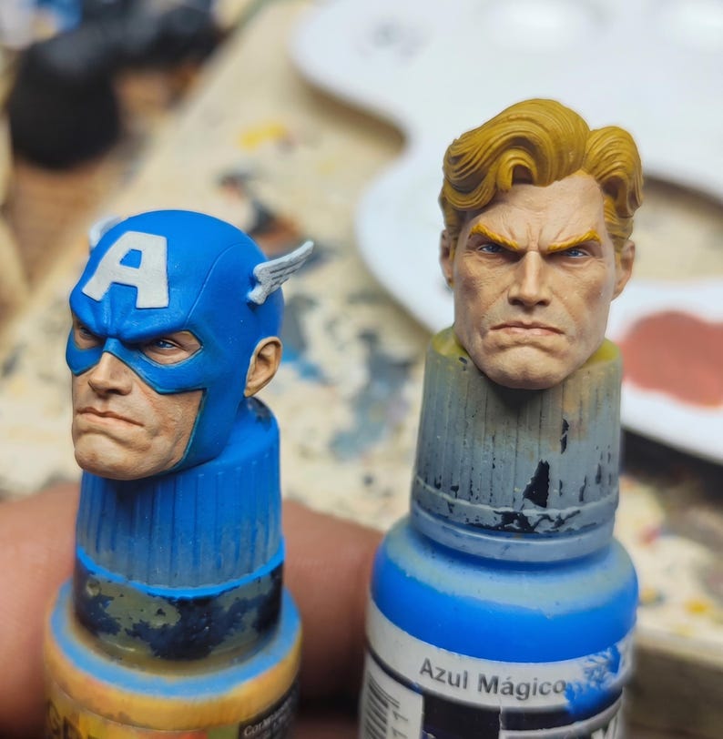 May include: Two miniature painted heads mounted on paint bottles. One head features a blue mask with a white letter A and small wings, while the other head shows a person with blonde hair and a serious expression. Both have detailed facial features.