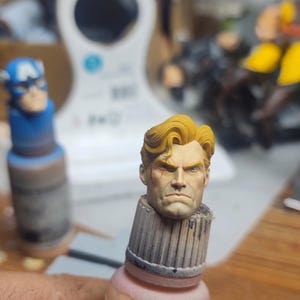 May include: A miniature hand-painted head sculpture featuring a male figure with blonde hair, a stern facial expression, and a small red mark on the cheek. The head is mounted on a cylindrical base, positioned next to a blue Captain America figure.