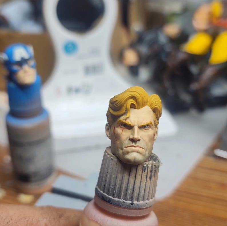 May include: A miniature hand-painted head sculpture featuring a male figure with blonde hair, a stern facial expression, and a small red mark on the cheek. The head is mounted on a cylindrical base, positioned next to a blue Captain America figure.