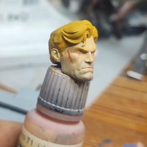 May include: A miniature painted head with blonde hair and a visible red scar on the right cheek, mounted on top of a paint bottle cap. The head features detailed facial expressions, defined eyebrows, and a serious, focused look on the face.