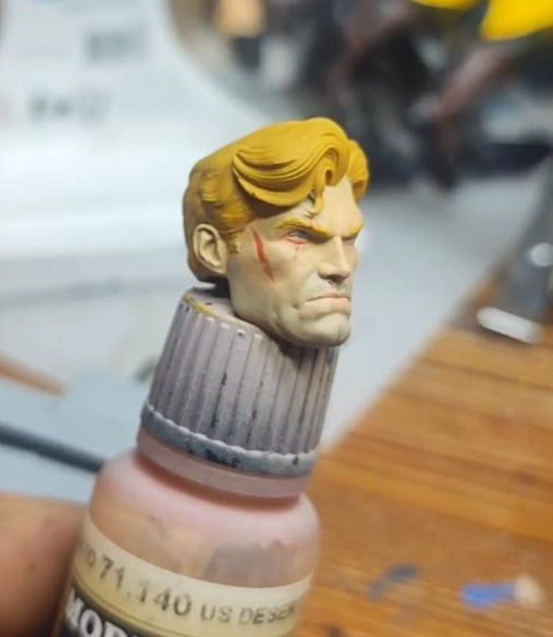 May include: A miniature painted head with blonde hair and a visible red scar on the right cheek, mounted on top of a paint bottle cap. The head features detailed facial expressions, defined eyebrows, and a serious, focused look on the face.