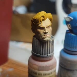 May include: A miniature painted head of a male figure with blonde hair and a stern facial expression, mounted on top of a small paint bottle labeled 340 US DESERT. The figure features detailed facial sculpting and a light skin tone.