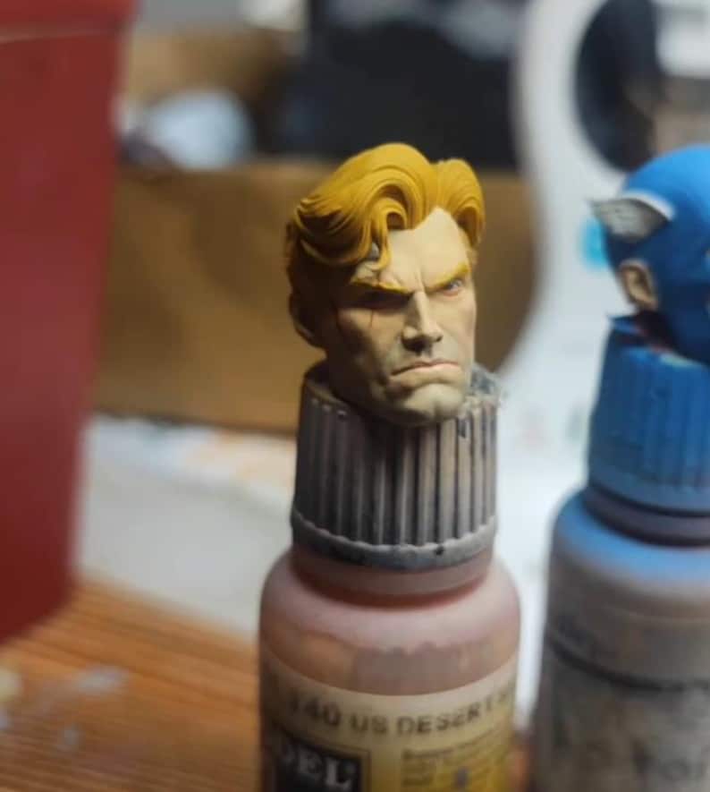 May include: A miniature painted head of a male figure with blonde hair and a stern facial expression, mounted on top of a small paint bottle labeled 340 US DESERT. The figure features detailed facial sculpting and a light skin tone.