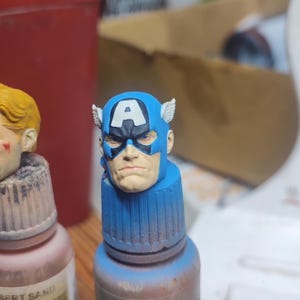 May include: A miniature painted head of Captain America featuring a blue cowl with a white letter A on the forehead and small white wings on the sides. The figure has a neutral facial expression with blue eyes and is mounted on a blue paint bottle cap.