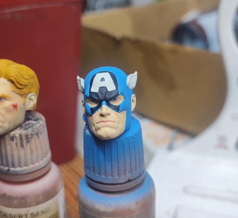 May include: A miniature painted head of Captain America featuring a blue cowl with a white letter A on the forehead and small white wings on the sides. The figure has a neutral facial expression with blue eyes and is mounted on a blue paint bottle cap.