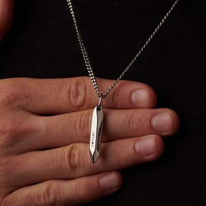 May include: A silver pendant necklace with a geometric design. The pendant is a long, slender shape with a faceted surface. The necklace is worn against a black shirt.
