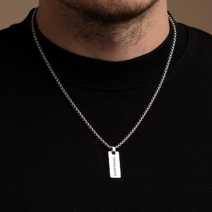 Könnte beinhalten: Eine silberne Kettenkette mit einem rechteckigen Anhänger, auf dem "SANTENO" in schwarzen Buchstaben steht.
