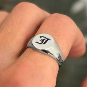 Op de afbeelding: Een gepolijste zilveren zegelring. De ring heeft een cirkelvormige voorkant met een zwarte, gestileerde letter "J". De ring wordt aan een vinger getoond.