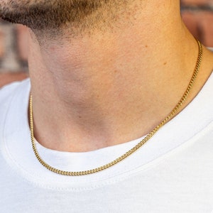 Könnte beinhalten: Eine goldene Kettenkette, die von einer Person mit einem weißen T-Shirt getragen wird. Die Kette ist im Curb-Stil und etwa 50 cm lang.