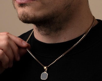 May include: A gold chain necklace with a small, square pendant. The pendant is a dark gray color with a gold border.