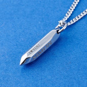 Puede incluir: Un collar con colgante plateado con un dije en forma de bala grabado con el nombre "SIMON" y las palabras "I YOU". La cadena es de diseño delicado y eslabonado. El colgante destaca sobre un fondo azul, resaltando el metal pulido.