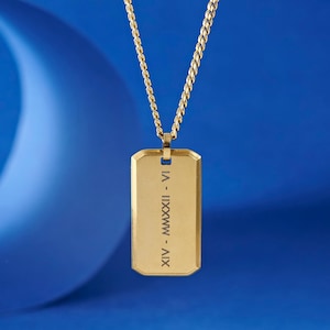 Könnte beinhalten: Ein goldfarbener Anhänger in Form einer Erkennungsmarke mit Kette, vor blauem Hintergrund. Die Marke hat eingravierte römische Ziffern. Die Kette ist im Curb-Stil gehalten. Der Anhänger ist rechteckig mit abgerundeten Ecken.