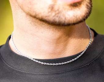 May include: A silver rope chain necklace on a person's neck. The chain is thin and has a delicate, twisted design. The person is wearing a black t-shirt.