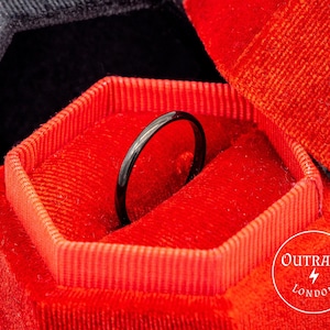 Puede incluir: Un anillo negro se encuentra dentro de una caja de anillo de terciopelo rojo. La caja es hexagonal y tiene el texto "OUTRAGE LONDON" en la parte inferior.