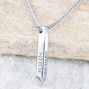 May include: A silver-colored pendant necklace with a polished, rectangular design. The pendant is engraved with the names "JOHN" and "RICHARD" in a vertical arrangement. The necklace chain is also silver and appears to be a curb chain style.