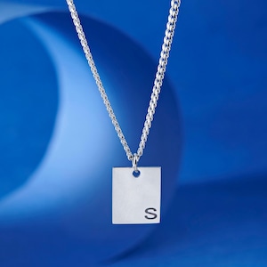 Pode incluir: Um colar prateado com um pingente quadrado com a letra "S". O pingente pende de uma corrente fina. O fundo é um gradiente de azul. Uma joia elegante.