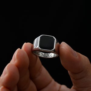 Peut inclure: Une bague chevalière argentée dotée d'une incrustation noire carrée et lisse sur le dessus. L'anneau intérieur de la bague est gravé des chiffres romains CMXC, clairement visibles sur la surface métallique polie de ce bijou.