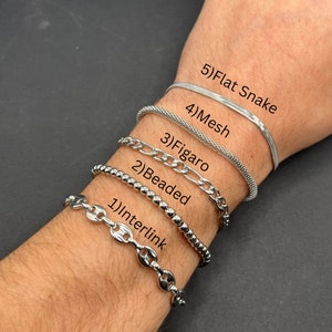 May include: Five different silver chain bracelets stacked on a person's wrist. The bracelets are labelled from top to bottom: Flat Snake, Mesh, Figaro, Beaded, and Interlink.