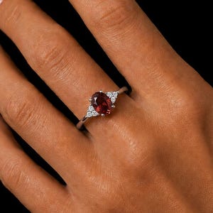 May include: A silver ring featuring a central oval-cut deep red gemstone flanked by two small triangular clusters of three clear white stones on each side, set on a thin polished metal band against a solid black background.