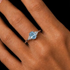 May include: A silver-toned ring featuring a central oval-cut light blue gemstone flanked by three small clear stones on each side. The band is thin and metallic, showcasing a polished finish that reflects light against the dark background.