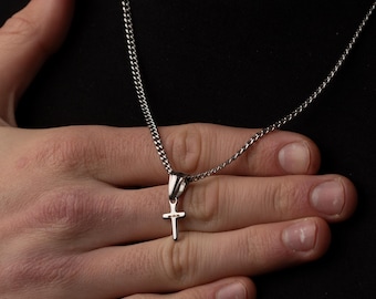 May include: A silver-colored cross pendant necklace. The cross is small and hangs from a delicate chain. The necklace is displayed against a dark background.