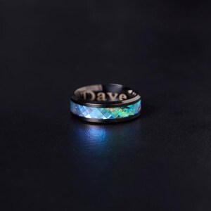 Könnte beinhalten: Ein schwarzer Ring mit dem eingravierten Namen "Dave" auf dem Band. Der Ring hat eine facettierte, irisierende Einlage mit Blau- und Grüntönen, die einen schimmernden Effekt erzeugt. Der Ring ist vor einem dunklen Hintergrund platziert.