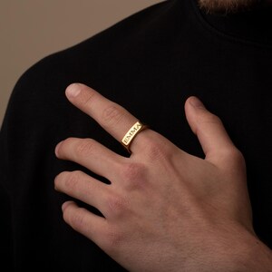 Könnte beinhalten: Ein goldfarbener Ring mit dem eingravierten Namen "EMMA" auf der Vorderseite. Der Ring ist rechteckig und wird an einem Finger getragen. Der Hintergrund ist eine Person, die ein schwarzes Oberteil trägt.