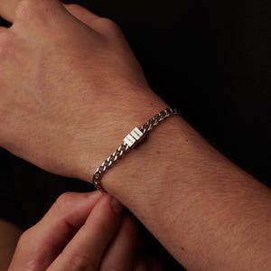 May include: A silver curb chain bracelet with a rectangular clasp. The clasp has the engraved word "LUCY" on it. The bracelet is on a person's wrist.