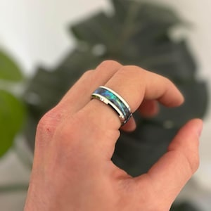 May include: A silver ring featuring a band of iridescent blue and green abalone shell. The ring is on a finger, highlighting the contrasting colours of the metal and the shell. The ring is a piece of jewellery.