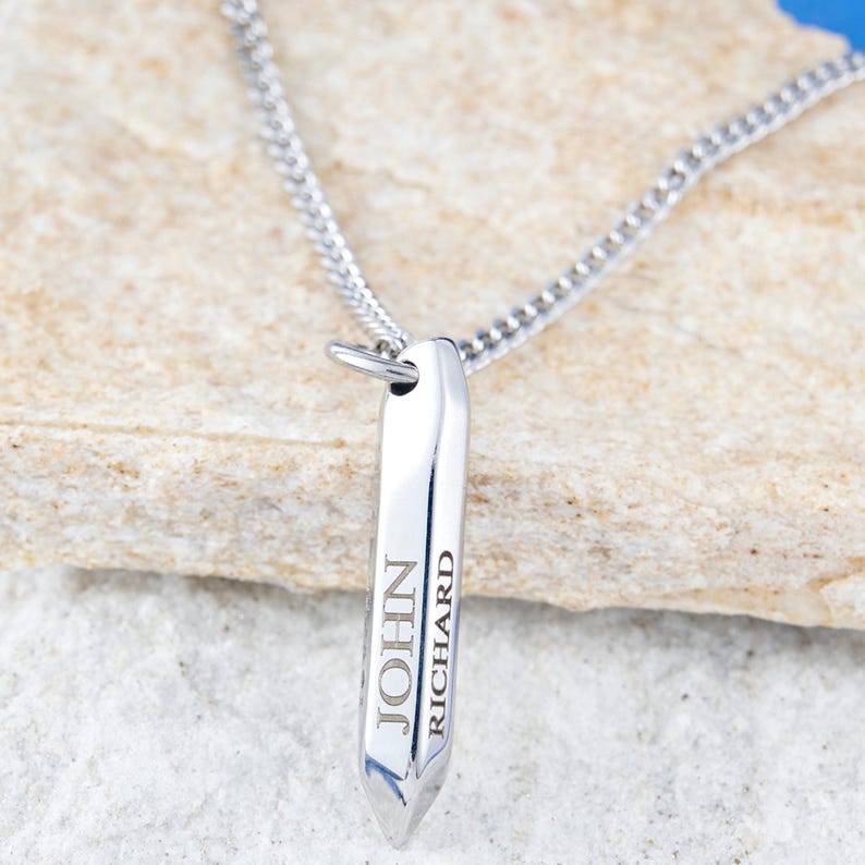 May include: A silver-colored pendant necklace with a polished, rectangular design. The pendant is engraved with the names "JOHN" and "RICHARD" in a vertical arrangement. The necklace chain is also silver and appears to be a curb chain style.