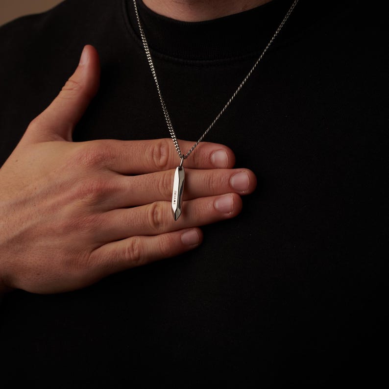 May include: A silver pendant necklace with a geometric design. The pendant is a long, slender shape with a faceted surface. The necklace is worn against a black shirt.