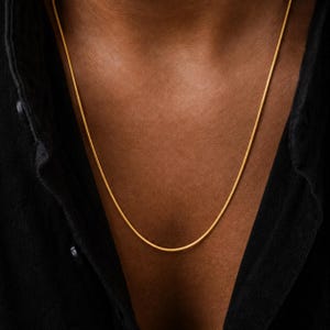 May include: A thin, smooth gold chain necklace resting against the skin. The necklace features a simple, continuous link design with a polished metallic finish. It is worn with a black collared shirt that is partially open at the neckline.