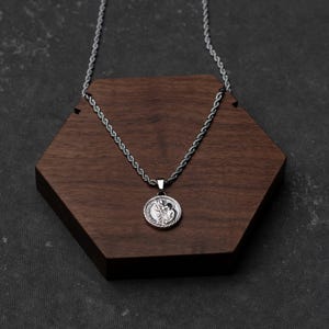 Puede incluir: Collar con colgante plateado con un dije circular detallado que representa una figura religiosa. La cadena tiene un diseño de cuerda. El colgante descansa sobre una base de madera hexagonal marrón oscuro.