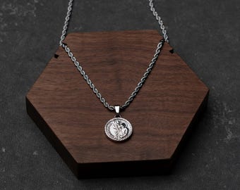 May include: A silver-colored pendant necklace with a detailed circular charm featuring a religious figure. The necklace chain has a rope-like design. The pendant rests on a dark brown, hexagonal wooden display.