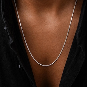 Puede incluir: Un collar de cadena de plata plano y fino tipo serpiente que descansa sobre el cuello. El collar presenta un acabado metálico liso con un diseño de eslabones entrelazados flexibles que crea una superficie continua, elegante y brillante.
