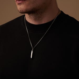 May include: A silver-colored pendant necklace with a small, elongated, pointed pendant. The pendant hangs from a delicate chain. The necklace is worn against a black shirt.