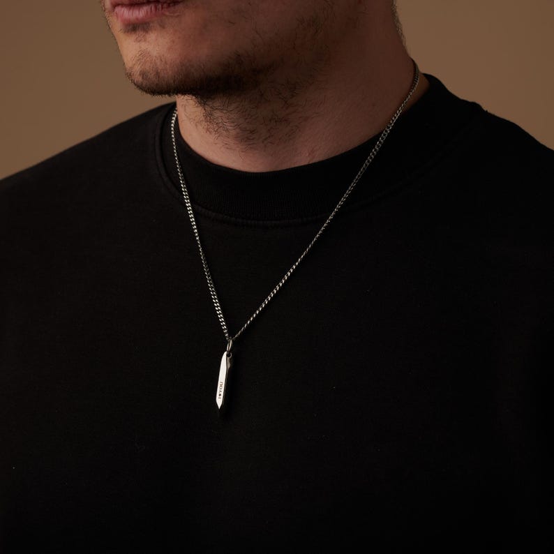 May include: A silver-colored pendant necklace with a small, elongated, pointed pendant. The pendant hangs from a delicate chain. The necklace is worn against a black shirt.