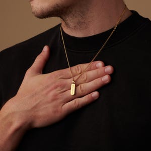 May include: A gold-colored necklace with a rectangular pendant. The pendant has the Roman numerals "MMXXIII" engraved on it. The necklace is worn against a black shirt.