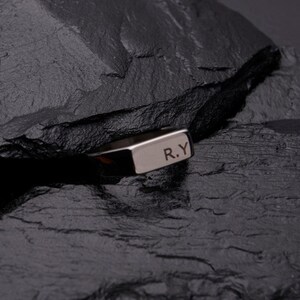 May include: A silver ring with the initials "R.Y" engraved on the top. The ring is rectangular and sits on a dark, textured surface. The ring is a simple, elegant piece of jewelry.