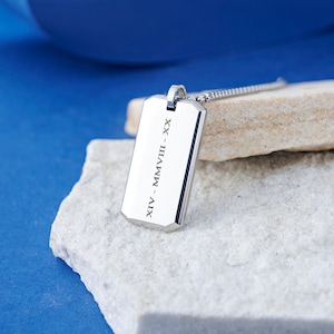 Könnte beinhalten: Silberner Anhänger im Dog-Tag-Stil mit Kette. Der rechteckige Anhänger hat abgeschrägte Kanten und die römischen Ziffern XIV - MMVIII - XX sind eingraviert. Der Anhänger liegt auf einem hellen Stein vor blauem Hintergrund.