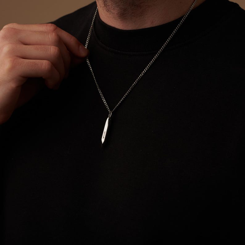 May include: A silver pendant necklace with a slender, elongated design. The pendant hangs from a delicate chain. The necklace is worn against a black shirt.