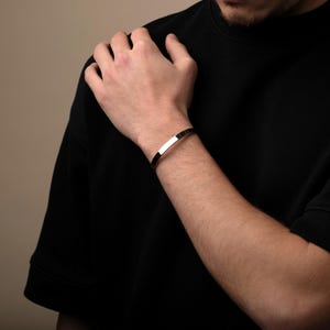 May include: A silver cuff bracelet with a flat, rectangular design. The bracelet is worn on the wrist and has a polished, reflective surface. The jewelry is a simple, modern accessory.