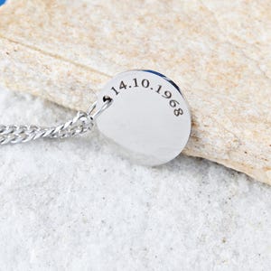 May include: A silver-colored pendant necklace with a circular pendant. The pendant has the engraved date "14.10.1968" and is attached to a chain. The necklace is displayed on a textured, light-colored surface.