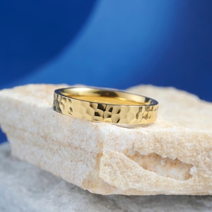 Puede incluir: Un anillo dorado con textura martillada descansa sobre una piedra clara sobre un fondo azul. El anillo tiene una banda con superficie texturizada. El anillo es de estilo banda simple.