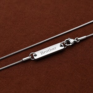 May include: A silver-toned snake chain necklace featuring a rectangular metal tag engraved with the word Brother. The necklace includes a lobster claw clasp attached to the tag, creating a secure closure for this metallic jewellery piece.